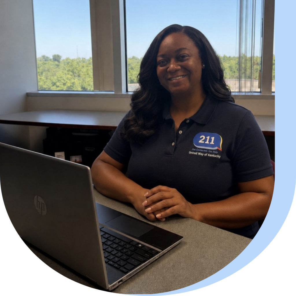 Smiling woman seated at a desk with an open laptop, wearing a navy polo with a 211 United Way of Kentucky logo, indoors by a window.